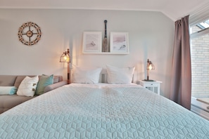 Una camera da letto con un letto grande, una sveglia a muro, due quadri incorniciati, un comodino con una lampada, una finestra con tende e un divano con cuscini.