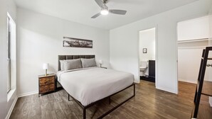 Ein Schlafzimmer mit einem Bett, einem Nachttisch, einem Deckenventilator, einem Stadtbild-Kunstwerk und einem durch eine offene Tür einsehbaren Badezimmer.