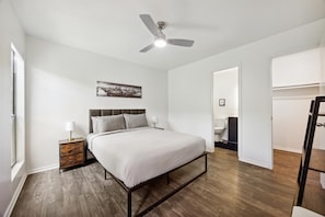Una camera da letto con un letto, un comodino, una ventola a soffitto, un'opera d'arte con una veduta sulla città e un bagno visibile attraverso una porta aperta.