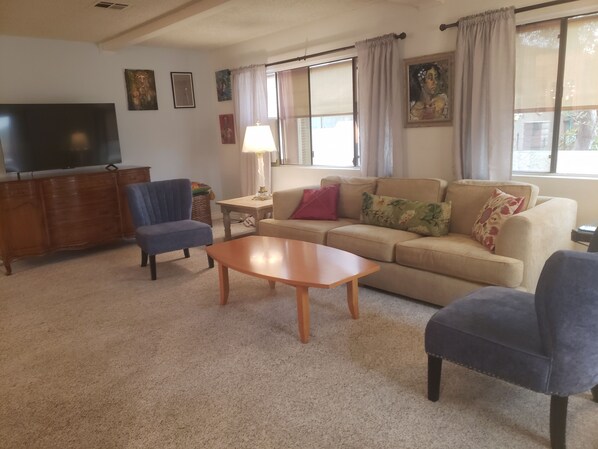 Una sala de estar con un sofá, dos sillones, una mesa de centro de madera, un televisor sobre un mueble de madera y una ventana con cortinas.
