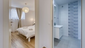 Un dormitorio con cama, mesita de noche y lámpara colgante. Incluye un baño con lavamanos y ducha. El cuarto tiene piso de madera y paredes blancas.