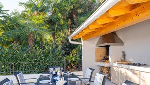 Un área de comedor al aire libre con una mesa larga dispuesta para seis personas, rodeada de sillas con cojines a rayas. Se ve un horno de ladrillo y una estufa a gas al fondo.