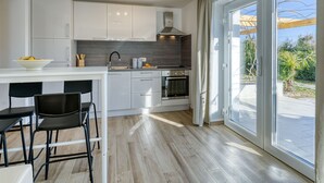 Una cocina moderna con armarios blancos, una isla central, taburetes negros y un amplio ventanal corredero que da a un espacio exterior.
