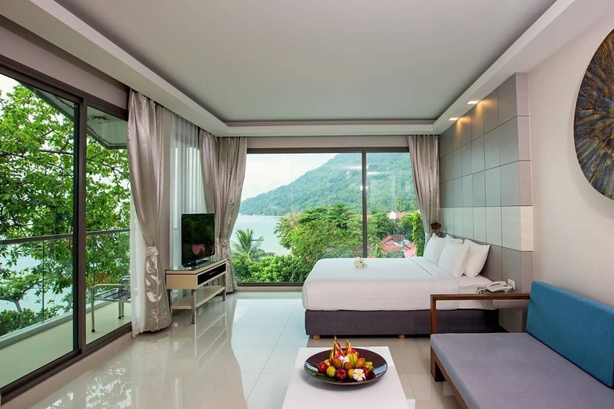 Ein modernes Hotelzimmer mit einem großen Bett, einem Fernseher, einem Esstisch mit Obst und Blick auf die Berge.