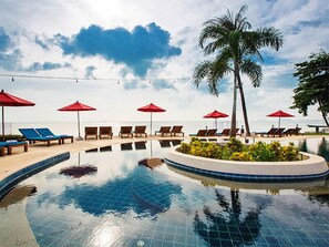 Zona de piscina con sillas de descanso y sombrillas rojas, rodeada de palmeras y vista al mar.