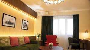A modern hotel room with a green sofa, a red armchair, a round wooden table, and a desk with a lamp. There are two framed pictures on the wall, a vase with flowers on a side table, and a large window with curtains. - Bayangol Hotel (Ulaanbaatar)
