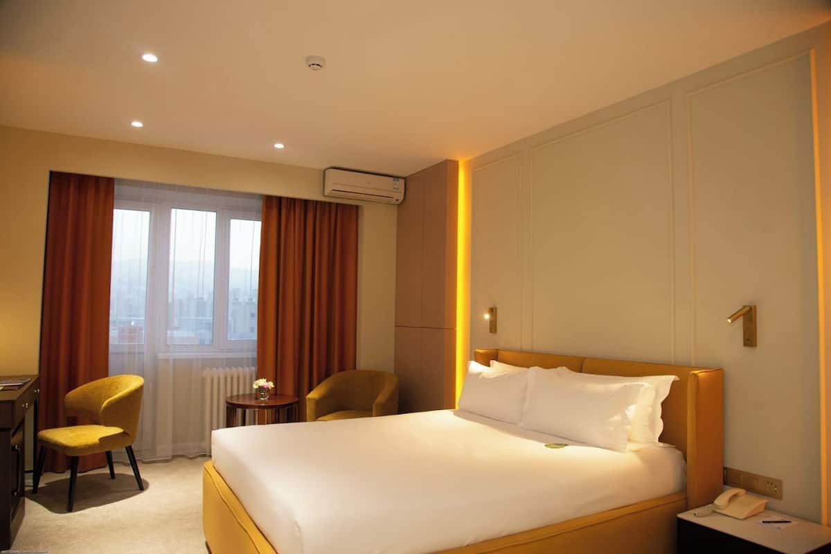 A hotel room with a bed, a desk, two chairs, a small table with a vase, and a window with curtains.