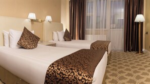 A hotel room with two beds, each with patterned bedspreads and matching pillows. There are bedside lamps and a floor lamp. The room has a large window with curtains. - Bayangol Hotel (Ulaanbaatar)