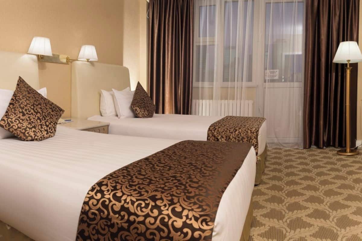 A hotel room with two beds, each with patterned bedspreads and matching pillows. There are bedside lamps and a floor lamp. The room has a large window with curtains.