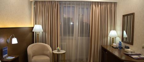 A hotel room with a large window, two lamps, a desk, a chair, and a small table with a vase of flowers.