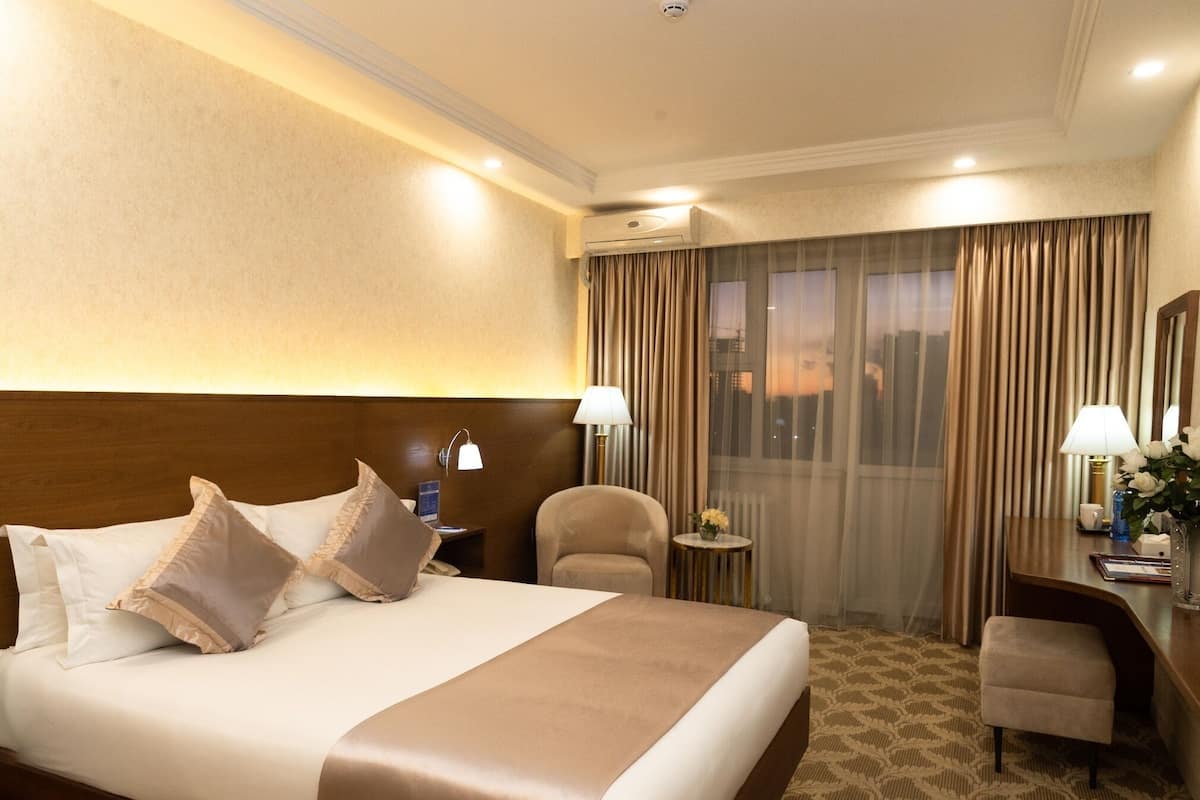 A hotel room with a large bed, a desk, a chair, and a view of the city through the window.