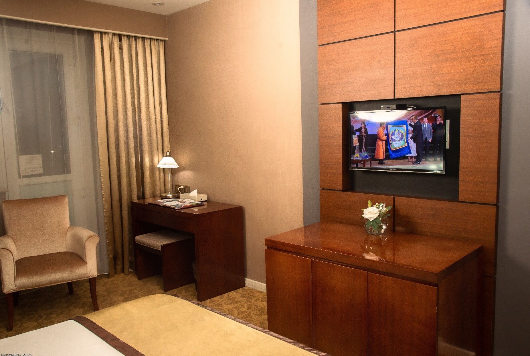 Habitació d'hotel amb televisió penjada a la paret, escriptori amb làmpada, una cadira i un petit armari amb un ram.