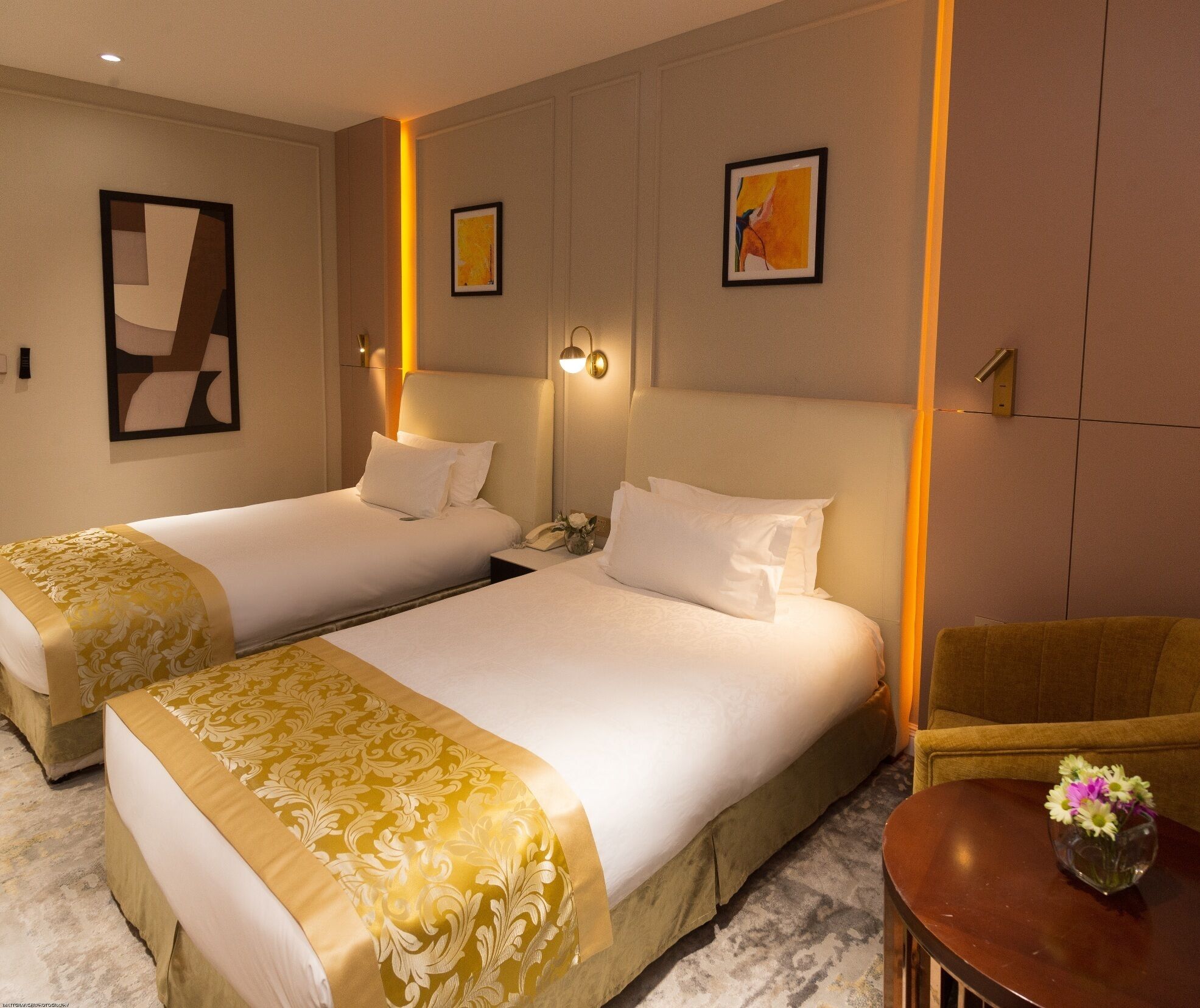A hotel room with two beds, a small table with a vase of flowers, and artwork on the walls.