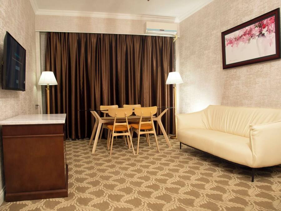 A hotel room with a beige sofa, a small table with chairs, a desk, and a large window with curtains.