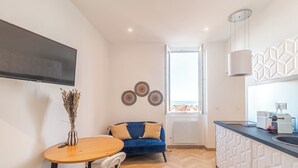 A modern kitchen with a built-in oven, a dining area with a wooden table and chairs, a blue sofa, and decorative wall art. - T2 Town Center, Casino (Beausoleil)