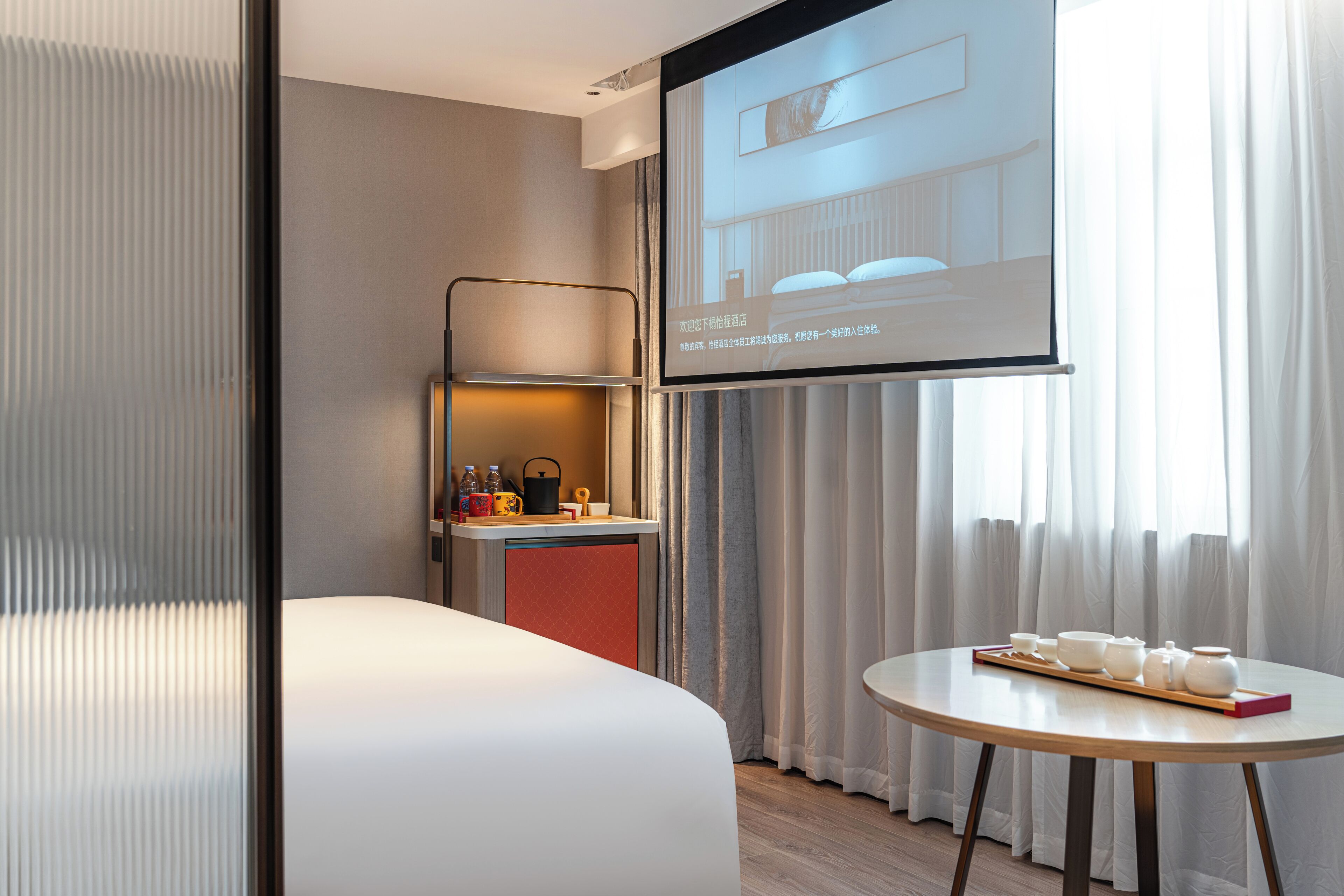 A hotel room with a bed, a small table with a tray of items, a mini-fridge, and a projection screen displaying a room image.