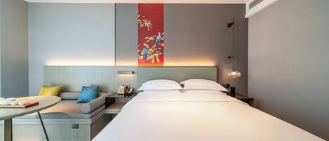 Una habitación de hotel moderna con una cama grande, una mesita de noche, un pequeño espacio para sentarse y una obra de arte colgada en la pared.