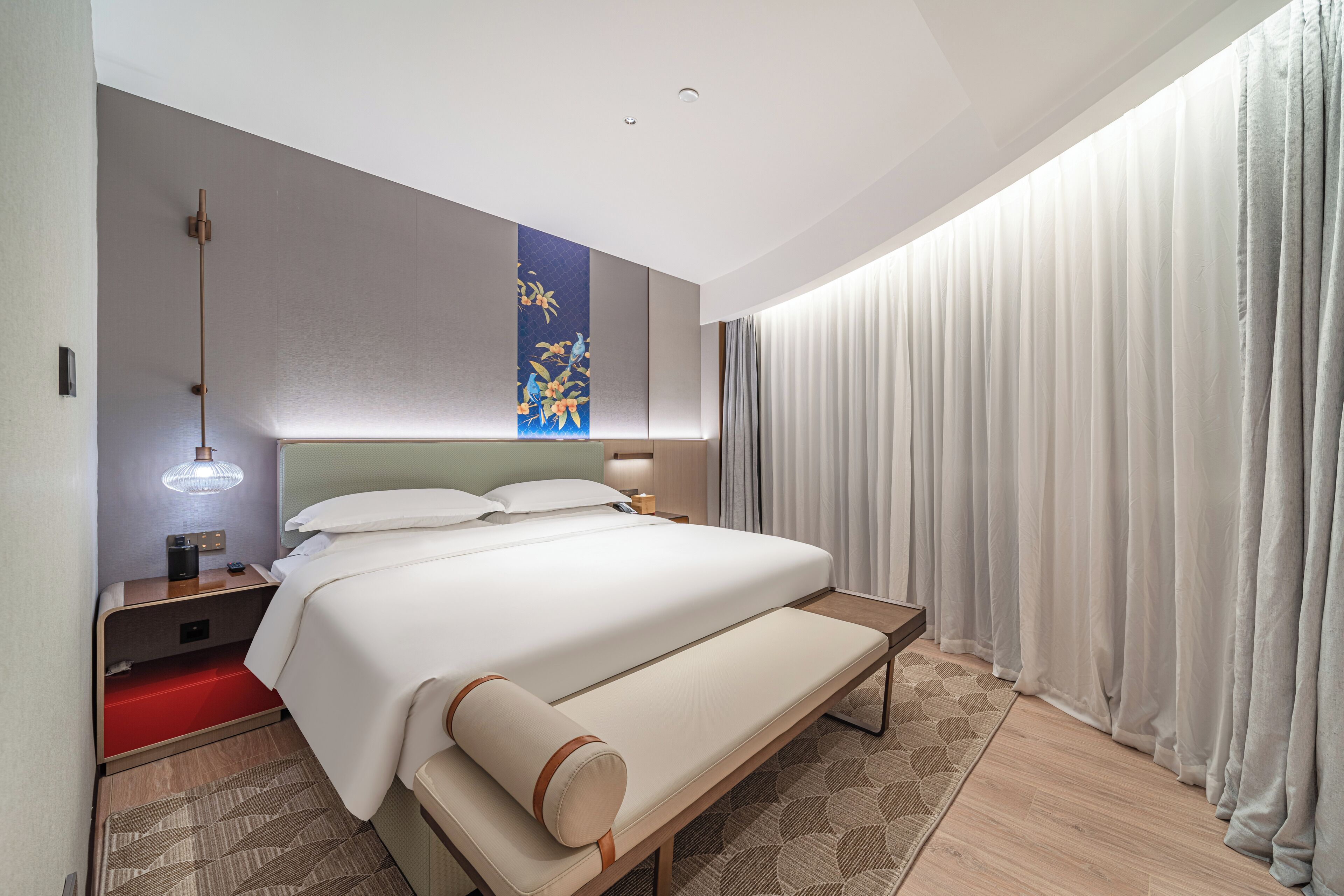 A modern hotel room with a large bed, a bedside table with a lamp, a wall-mounted artwork, and a view of the outside through sheer curtains.