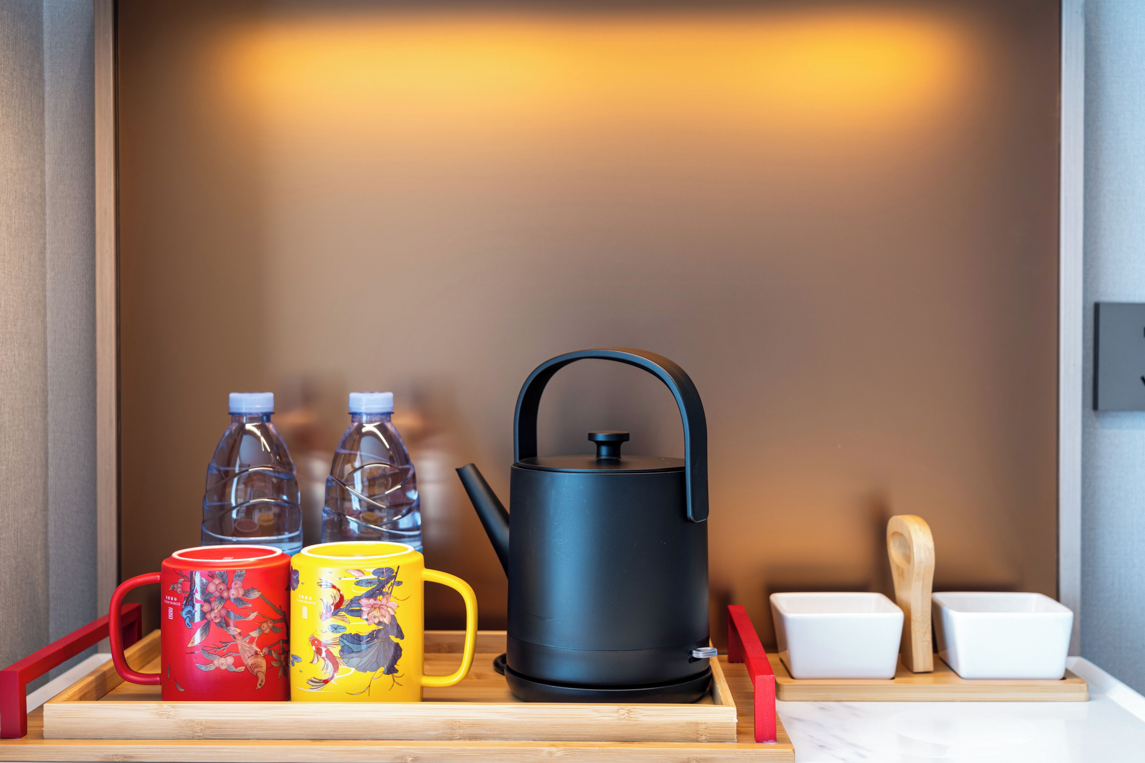 A kitchen area with a kettle, two water bottles, a red mug, and a yellow mug on a wooden tray.