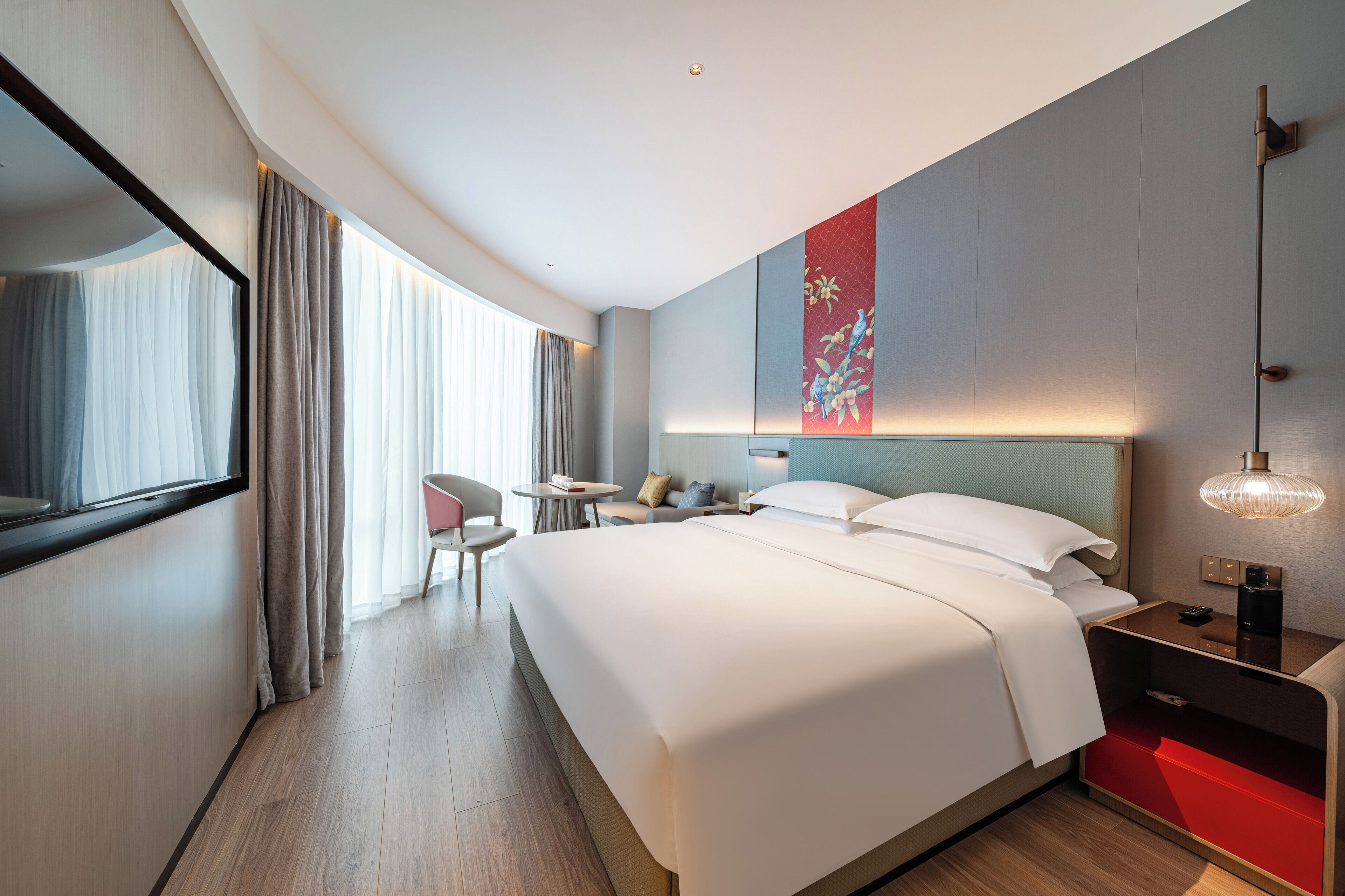 A modern hotel room with a large bed, a desk with a chair, a small table, and a red storage unit.