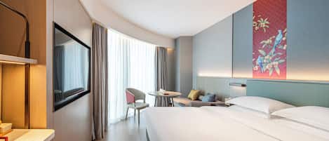 Habitación de hotel moderna con una cama grande, un escritorio, una silla y una mesita. Se aprecia una ventana con cortinas y un cuadro en la pared.