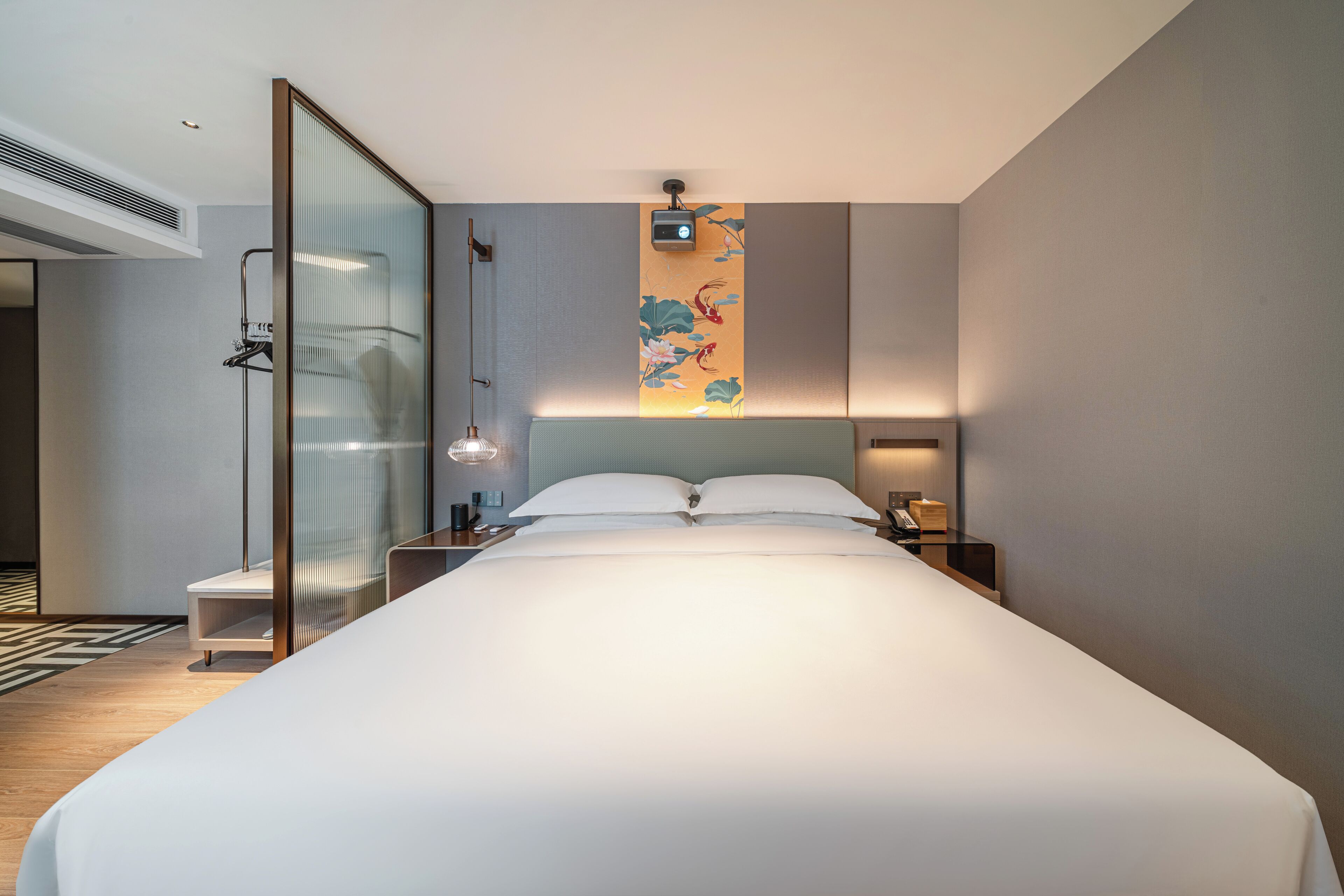 Een moderne hotelkamer met een groot bed, een glazen scheidingswand, een nachtkastje en een aan de muur bevestigde beamer.