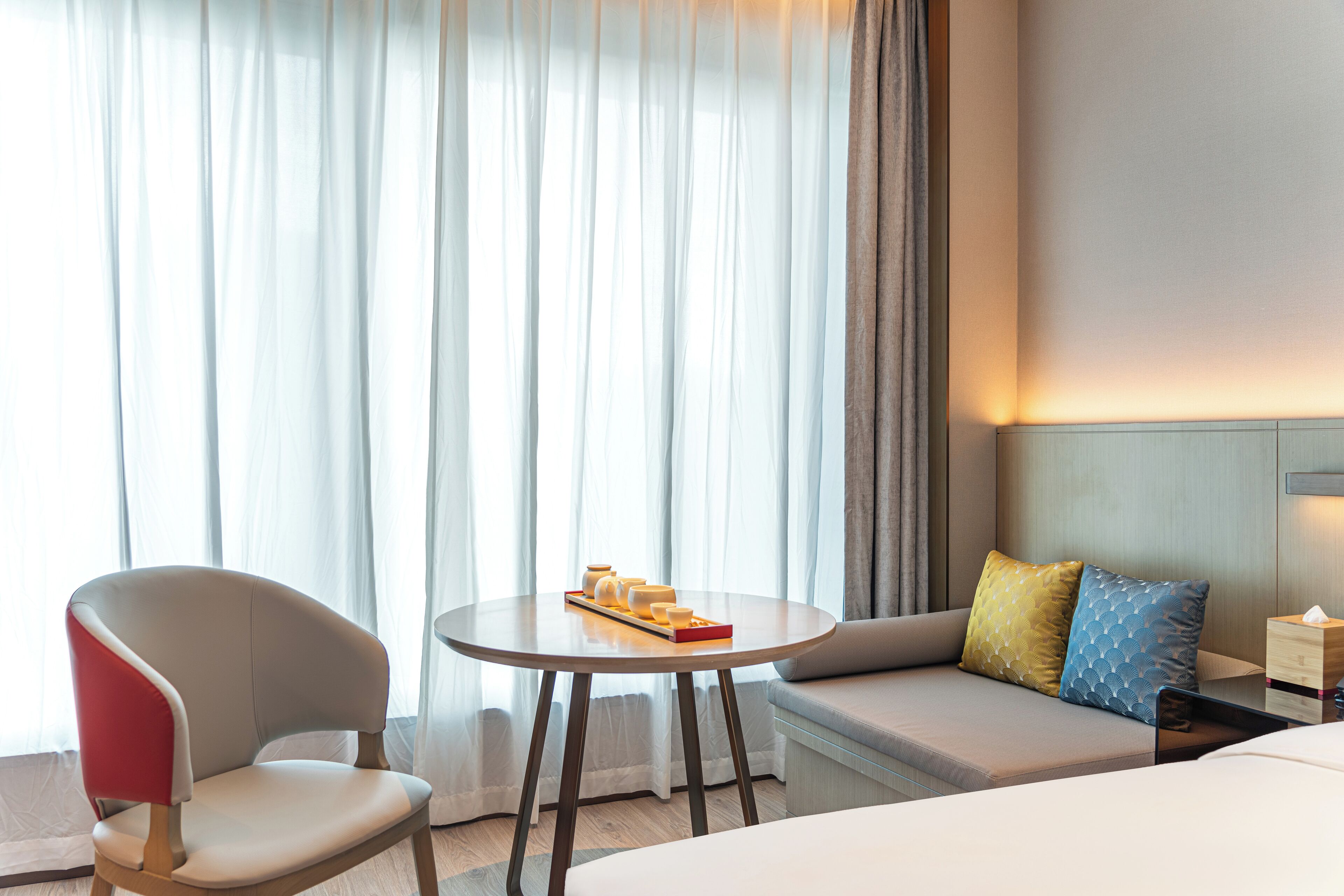 A hotel room with a sofa, a chair, a small table with a tray of items, and a bedside table with a box.