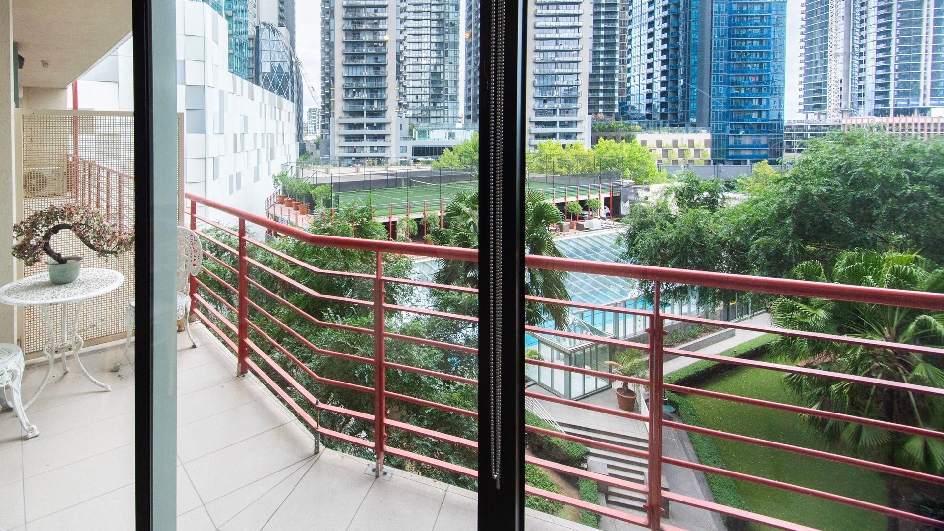 A balcony with a red railing overlooking a cityscape with modern buildings and a green park area.