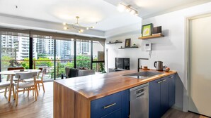 A modern kitchen with a wooden countertop, blue cabinets, and a dishwasher. There is a dining area with a table and chairs, and a large window offering a view of the city.