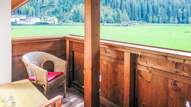 A wooden deck with a wicker chair and a table, overlooking a green field and forested hills. - Apart Jacqueline by Interhome (Huben)