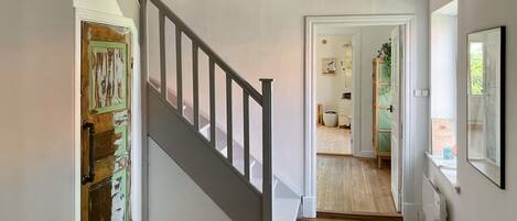 A staircase with a wooden handrail and a white wall. A door on the left is partially open, revealing a room with a green door. A framed picture hangs on the wall above the staircase.