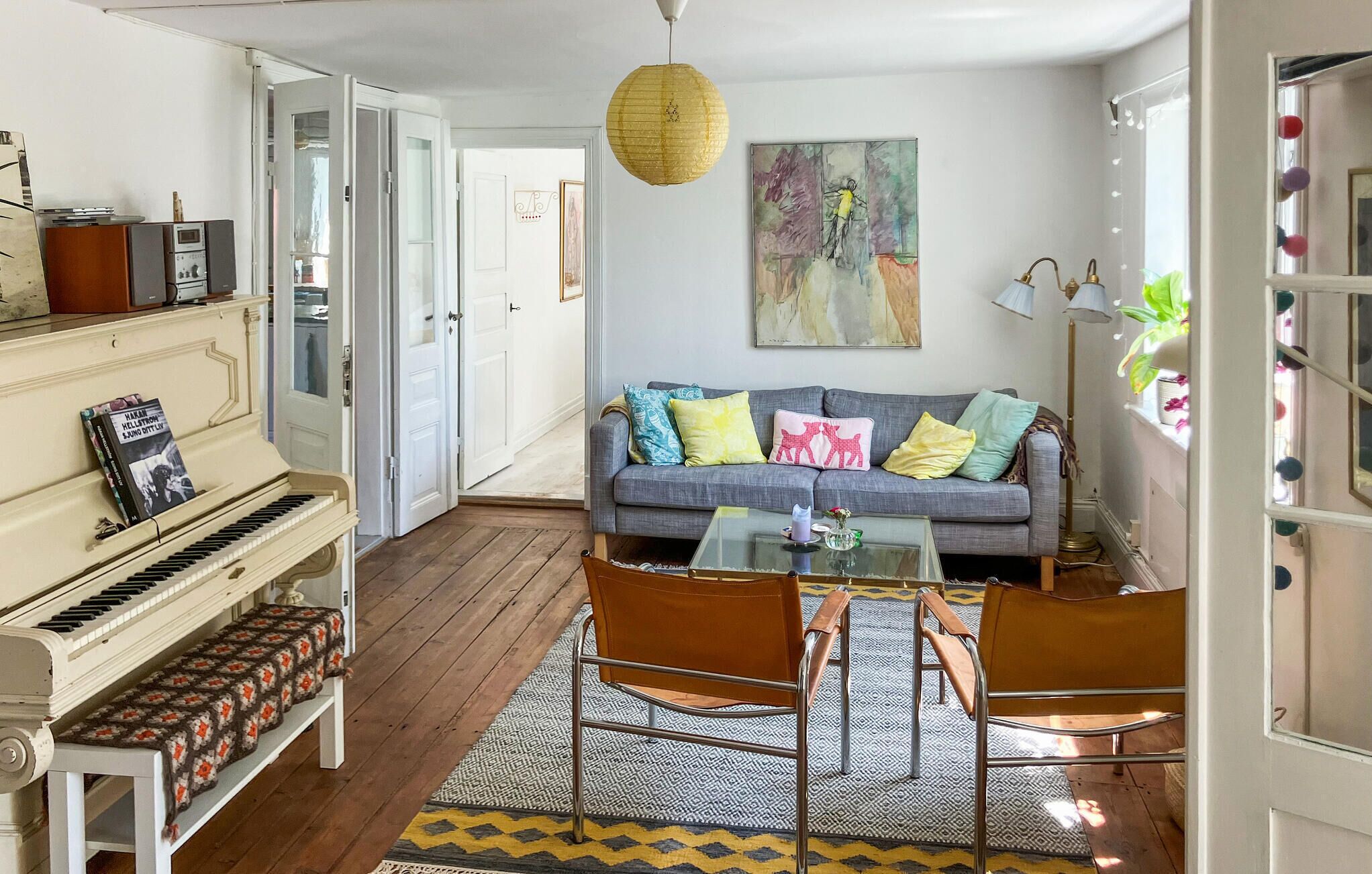 A living room with a piano, a sofa with colorful cushions, a glass coffee table, and a patterned rug.