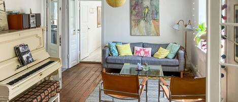 A living room with a piano, a sofa with colorful cushions, a glass coffee table, and a patterned rug.