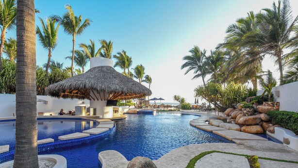 A resort pool area with a thatched-roof structure, palm trees, and a clear blue sky.