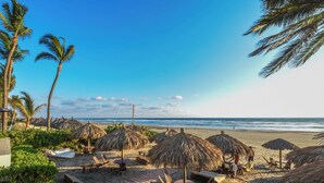 A beach with thatched umbrellas, lounge chairs, and palm trees.