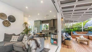 A modern living room with a grey sofa, a dining area, and a wooden deck with outdoor seating. - Barefoot Bay Cottage (Byron Bay)