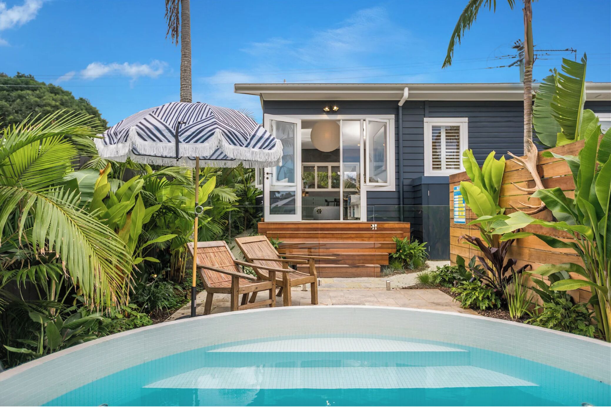 A poolside area with a pool, wooden deck, lounge chairs, and tropical plants.