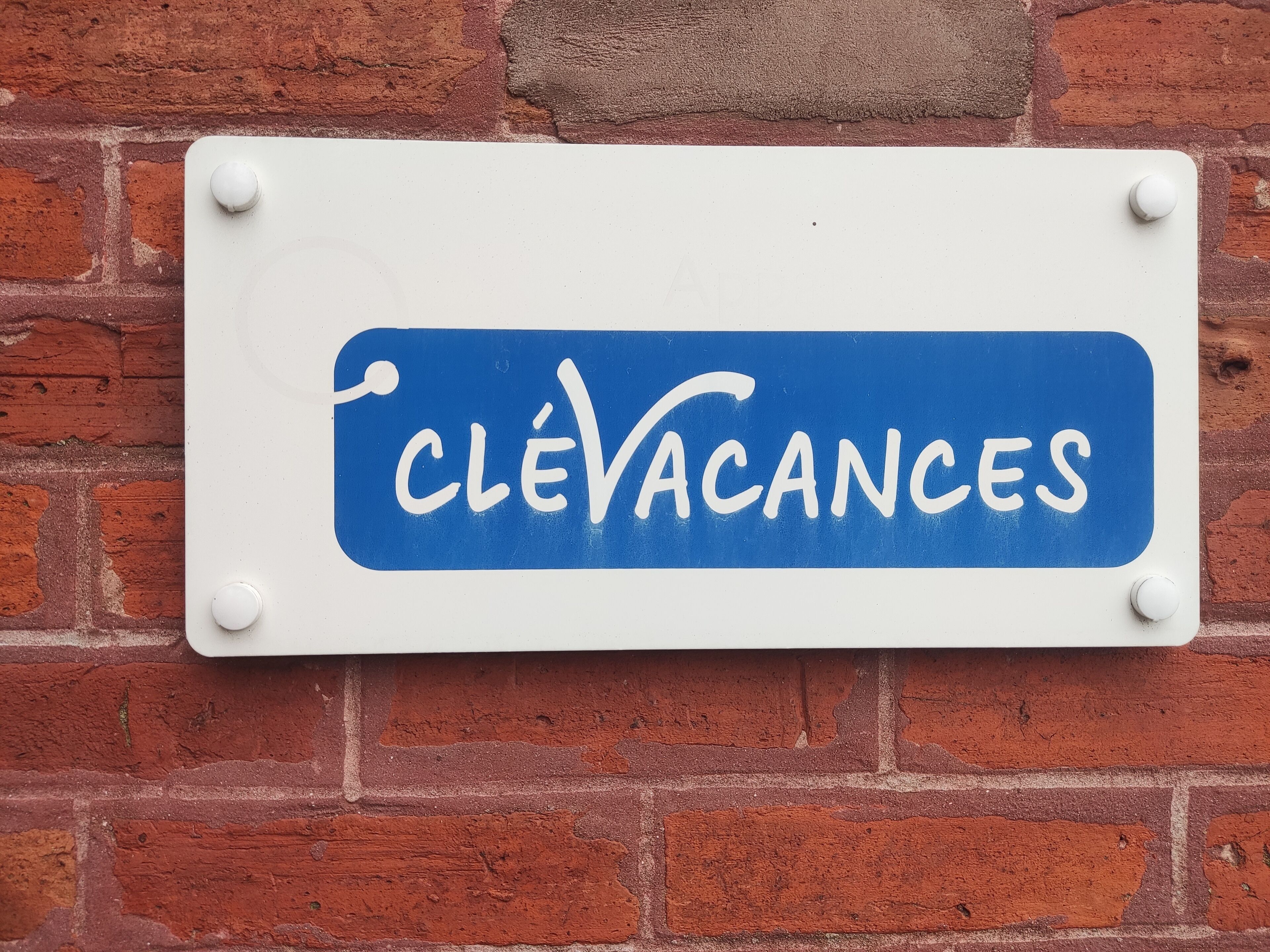 Ein Schild mit dem Text "CLÉVACANCES" vor weißem Hintergrund und blauer Umrandung, an einer Ziegelwand befestigt.