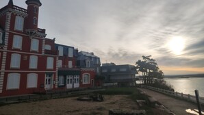 Un edificio histórico con una torre destacada y numerosas ventanas, ubicado frente a una ribera.
