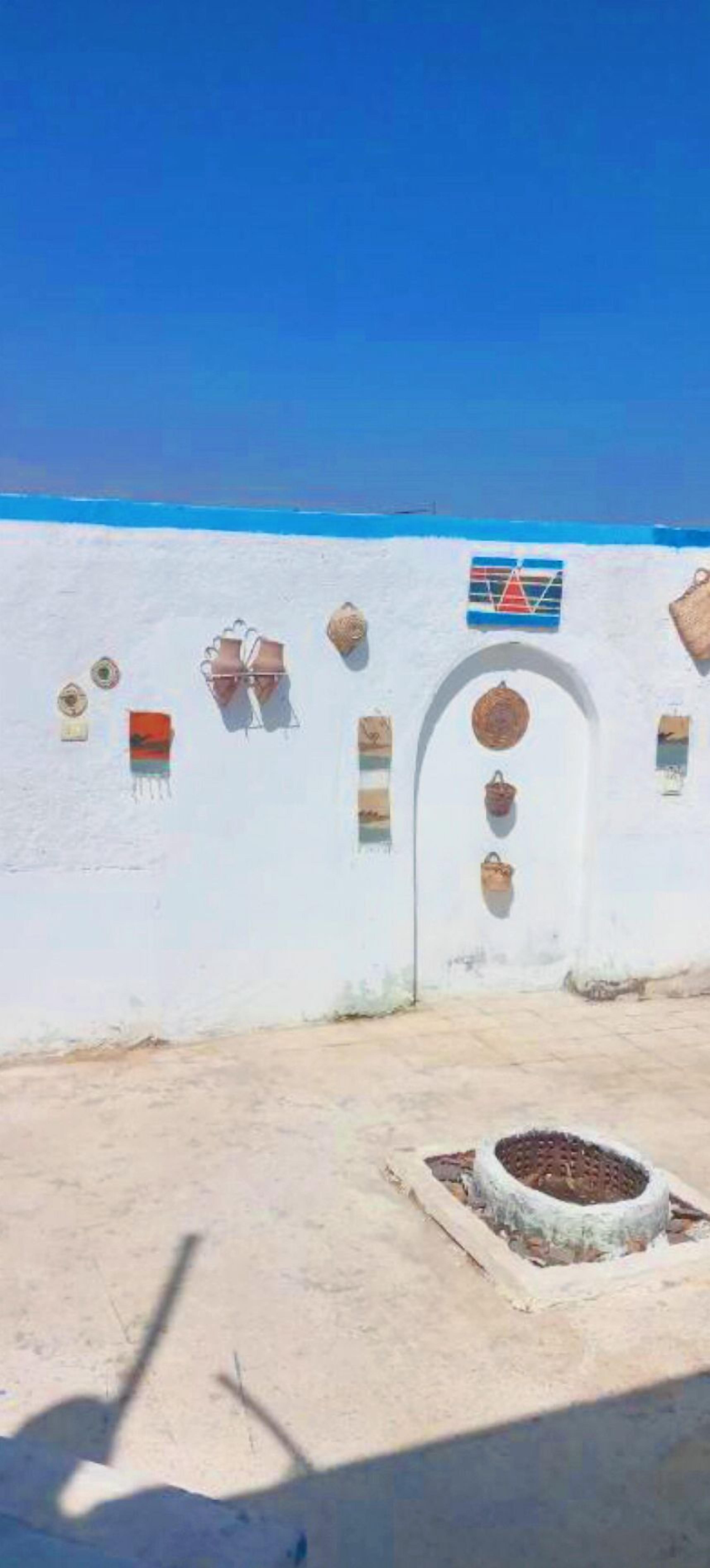Un bâtiment aux murs blancs, avec un ciel bleu en arrière-plan, une petite entrée et quelques éléments décoratifs, comme des paniers et un drapeau.