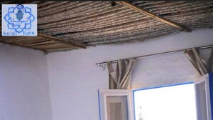 A room with a bed, a window with curtains, a wall with a blue and white pattern, and a thatched roof.