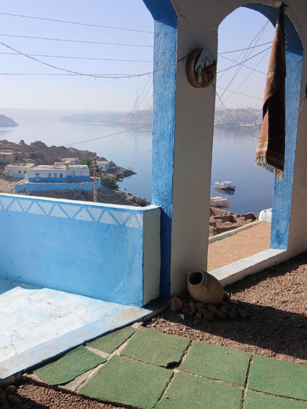Vista costera desde un balcón con pared pintada en azul y blanco, un pequeño espacio exterior con una maceta y una toalla, y una vista distante de barcos y edificios.