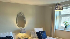 A hotel room with two beds, a bedside table with a lamp, a mirror, and a window with a view of greenery.