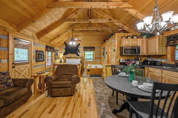 L’intérieur d’une cabane en rondins comprend une table à manger, des chaises, un canapé, un lit, un ventilateur de plafond, un lustre et un coin cuisine.