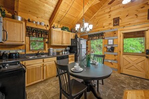 Une cuisine de style rustique, avec des éléments en bois, comprenant une table à manger, des chaises, un réfrigérateur, une machine à café et une fenêtre donnant sur un paysage verdoyant.