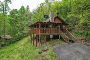 Une cabane en bois dotée d’une terrasse, d’un escalier et d’une cheminée, entourée d’une forêt dense.