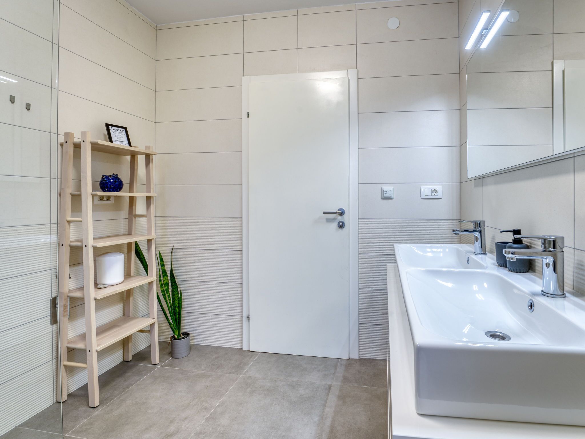 A modern bathroom with a white sink, a mirror, and a wooden shelf with decorative items.
