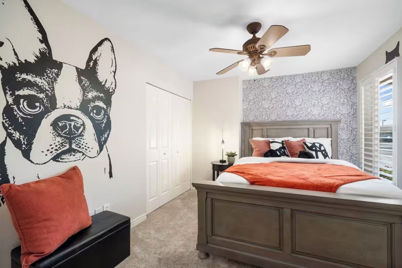 A bedroom with a large wall mural of a dog, a bed with a wooden headboard, a ceiling fan, and a nightstand with a lamp.