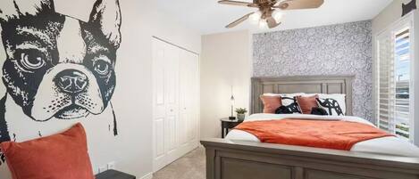 A bedroom with a large wall mural of a dog, a bed with a wooden headboard, a ceiling fan, and a nightstand with a lamp.