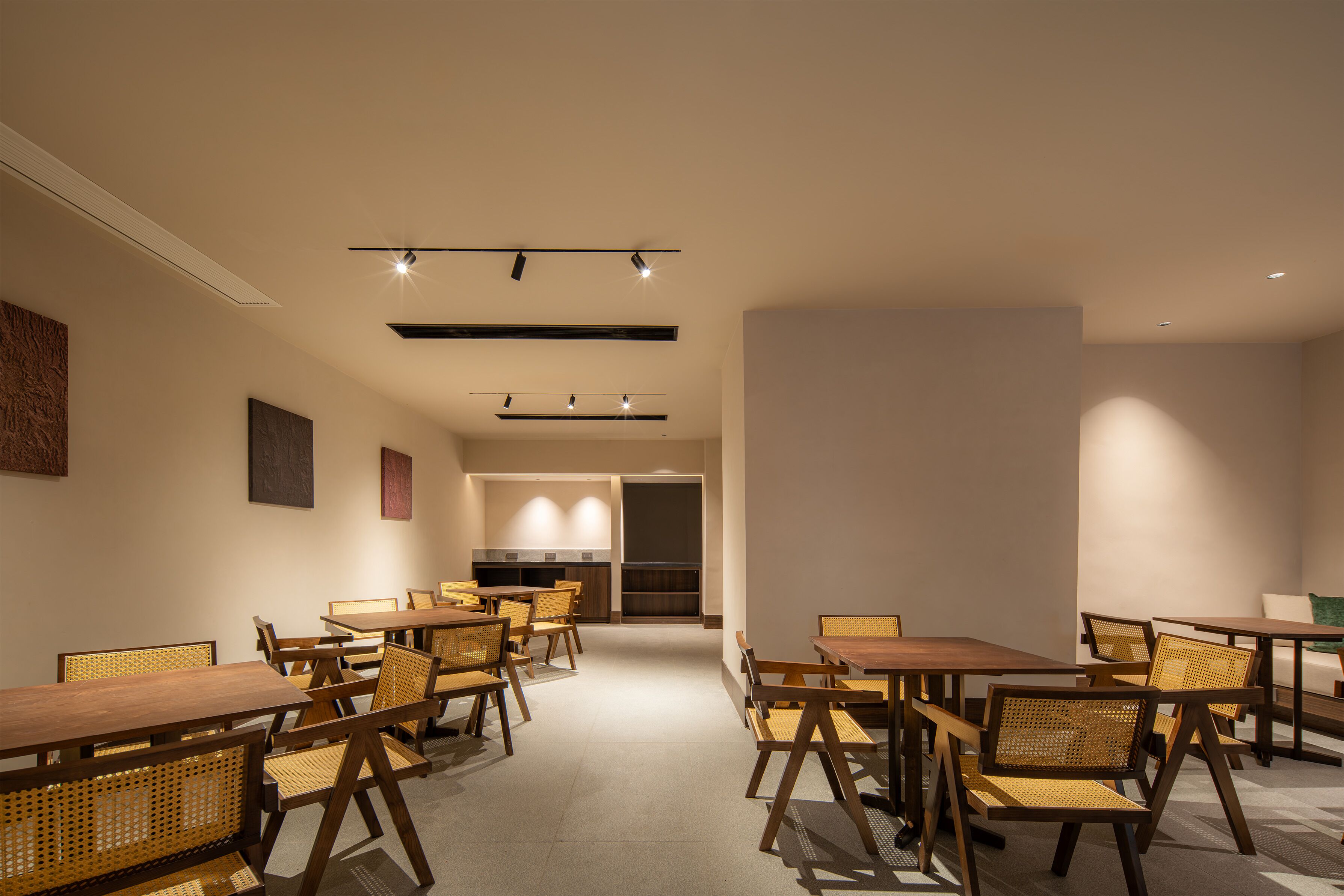 A modern dining area with wooden tables and chairs, recessed lighting, and a kitchen in the background.